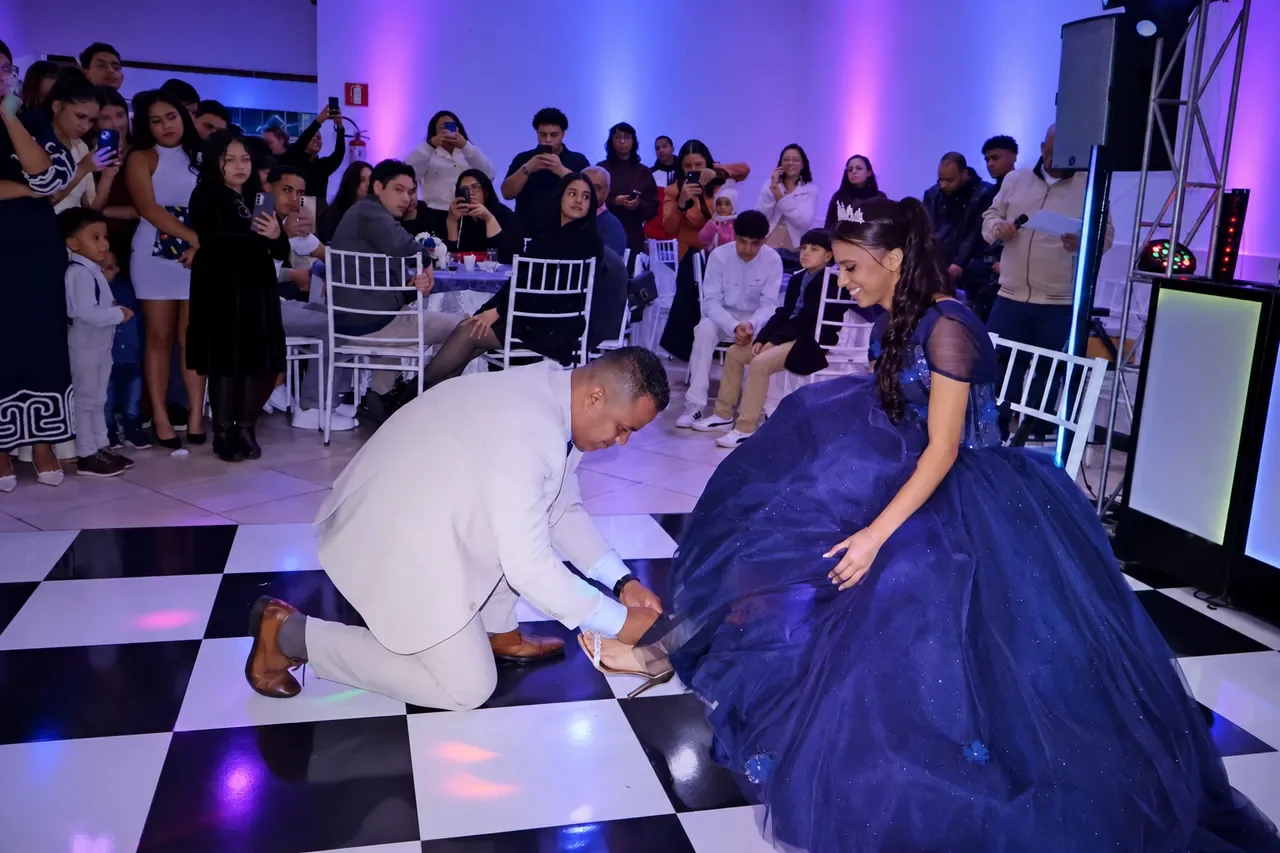 Alice sentada enquanto um familiar ajusta sua sandália durante a festa de 15 anos.