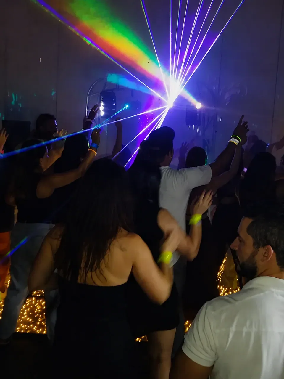 Celebração com pista cheia. Convidados dançando, braços levantados e iluminação colorida durante a celebração.